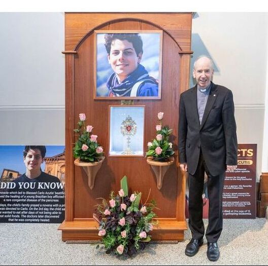 Carlo Acutis Ireland relic display with a portrait of Saint Carlo Acutis and a priest standing beside it