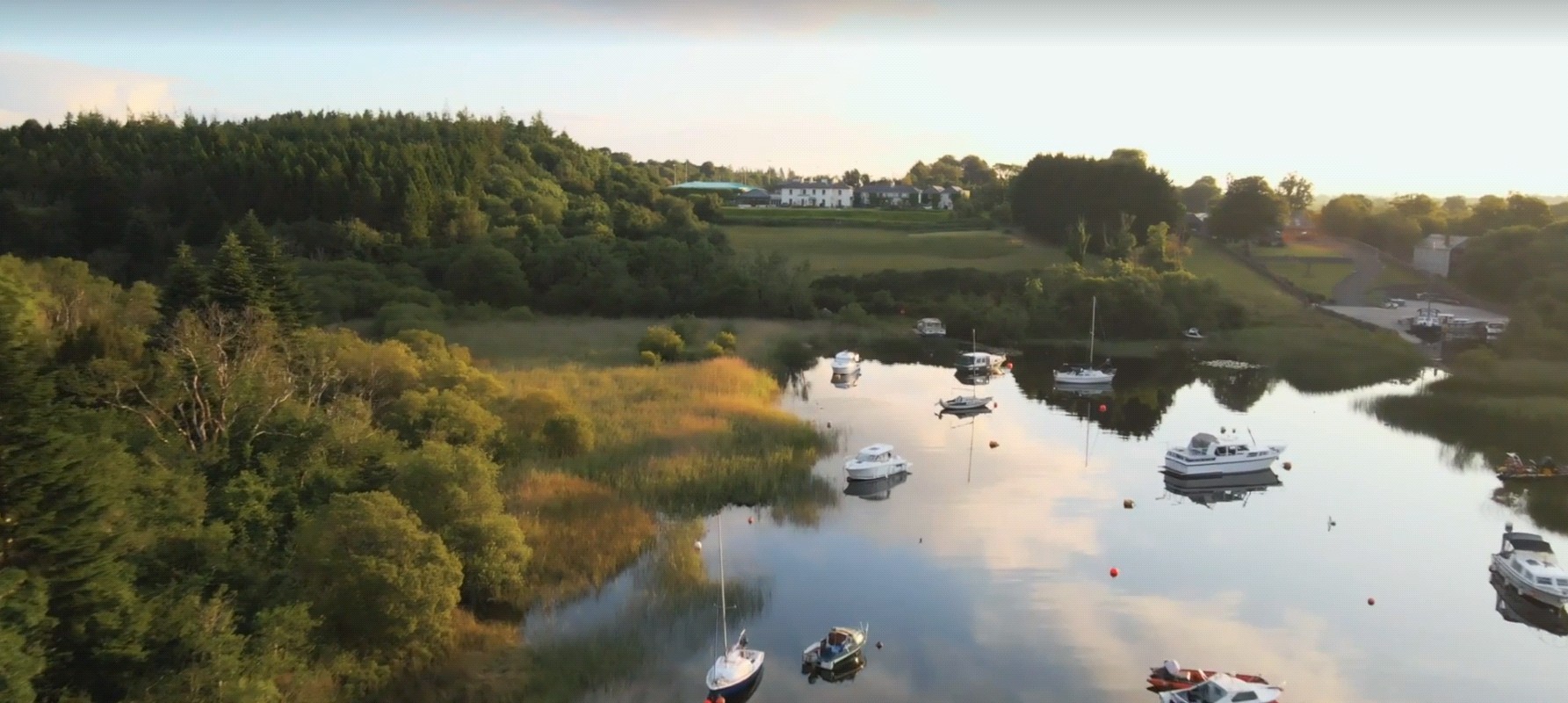 Aerial view looking down on the inlet of Lisloughrey and its harbour, with boats moored on calm water and woodland beyond
